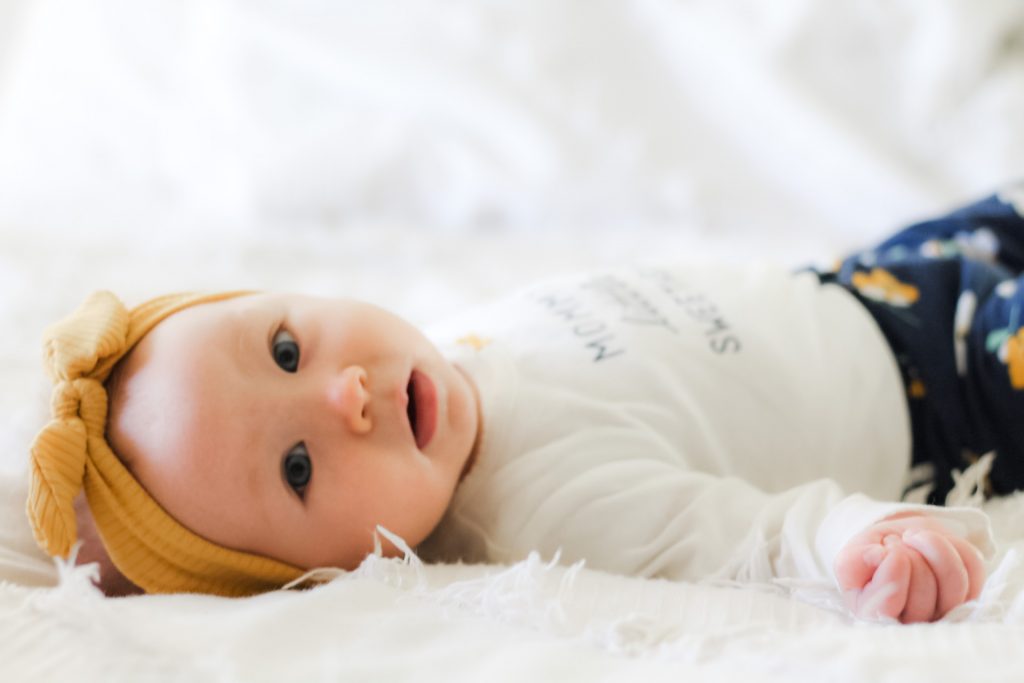 6 months baby girl with yellow bow laying on a white bedspread looking at camera